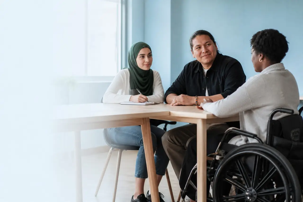 Trois personnes discutent autour d'une table : un professionnel autochtone, une collègue en fauteuil roulant et une jeune immigrante.