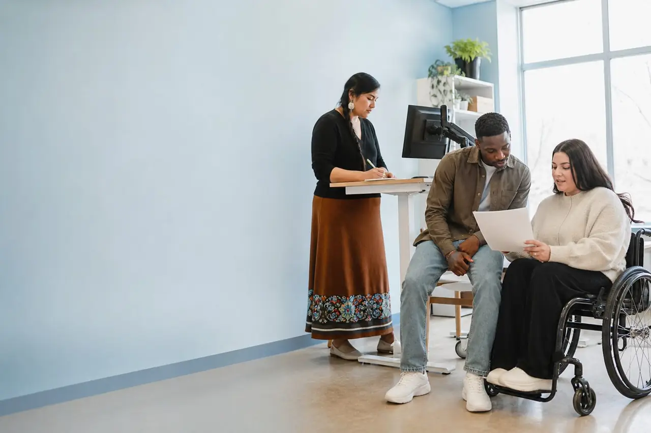 Une professionnelle autochtone rédige une offre pendant qu'un jeune immigrant et une collègue en fauteuil roulant relisent le brouillon.